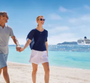 Ein Paar spaziert entspannt am sonnigen Sandstrand entlang, während im Hintergrund ein Kreuzfahrtschiff von Hapag-Lloyd Cruises im türkisblauen Meer ankert.