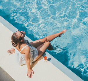 Frau sitzt entspannt am Rand eines Pools und hält die Füße ins Wasser, ein Getränk steht neben ihr.