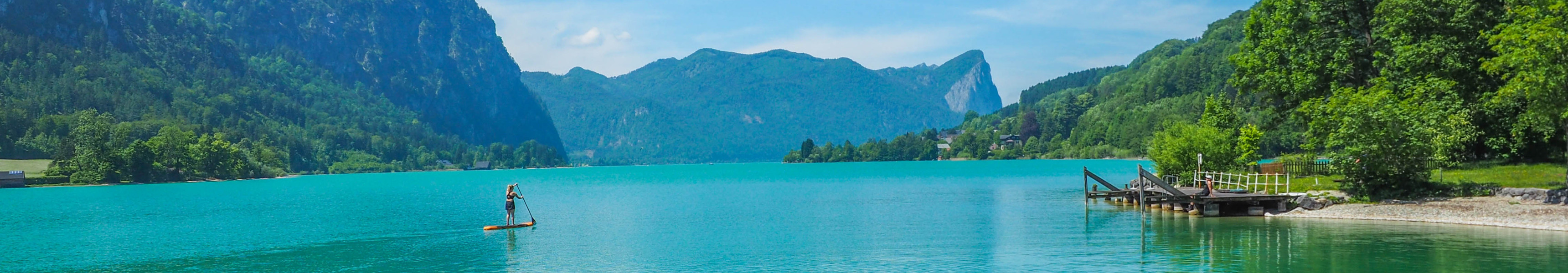 Panoramablick auf den t&uuml;rkisblauen Wolfgangsee in &Ouml;sterreich mit Bergen, Steg und Stand-up-Paddler