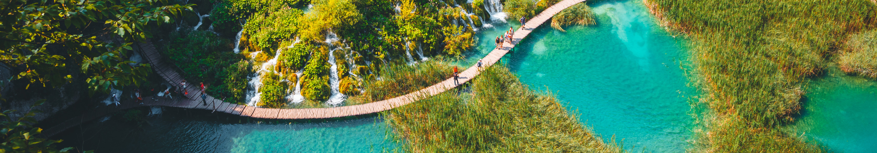 Holzstege f&uuml;hren durch die t&uuml;rkisfarbenen Seen und Wasserf&auml;lle des Nationalparks Plitvicer Seen in Kroatien, umgeben von gr&uuml;ner Vegetation und Felsen.
