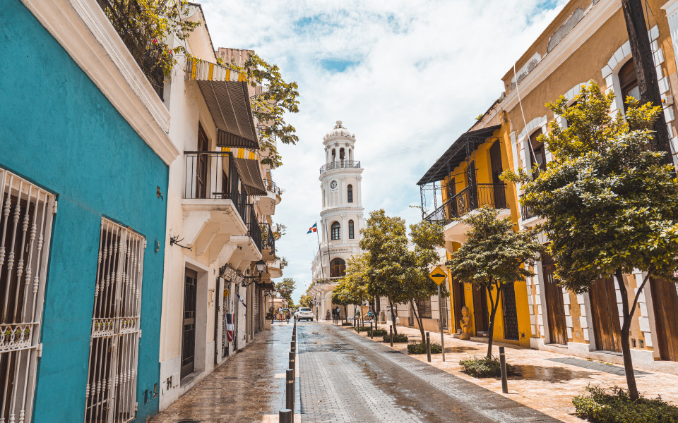 Stra&szlig;e mit bunten Kolonialh&auml;usern in Santo Domingo, Dominikanische Republik, mit einem wei&szlig;en historischen Turm im Hintergrund.