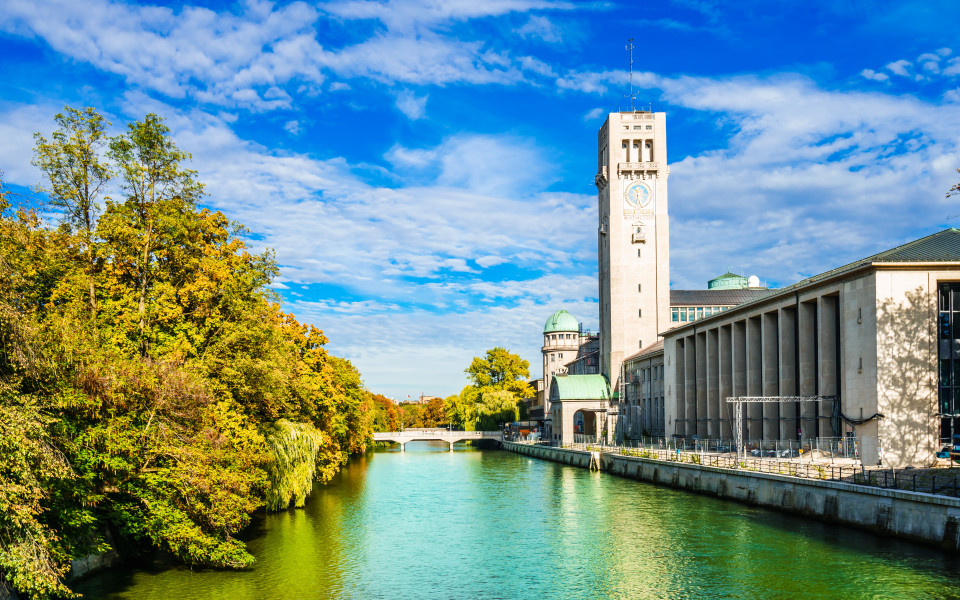 Sehensw&uuml;rdigkeit in M&uuml;nchen: Das Deutsche Museum in M&uuml;nchen, malerisch am Ufer der Isar gelegen, umgeben von herbstlich gef&auml;rbten B&auml;umen und strahlend blauem Himmel.