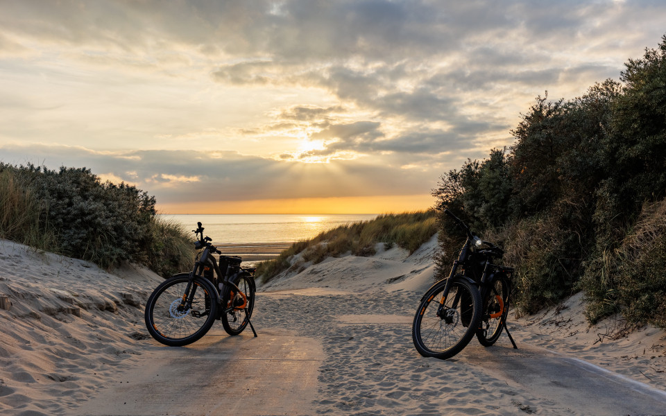 Zwei Fahrr&auml;der stehen in einer D&uuml;nenlandschaft auf einem sandigen Weg mit Blick auf das Meer und einen Sonnenuntergang am Horizont.
