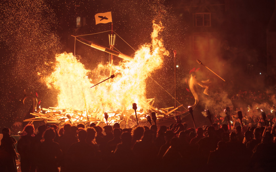 Ein großes, brennendes Wikingerschiff bei einem Feuerfest in der Nacht, mit vielen Zuschauern und Fackeln.